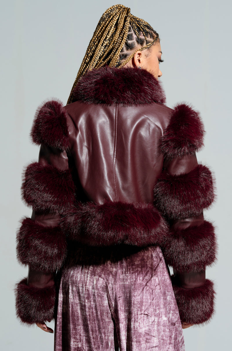 A person with braided hair is wearing the LOVERBOY Faux Fur Moto Jacket in burgundy, featuring a structured design with thick faux fur trim, along with purple textured pants, viewed from the back against a plain background.