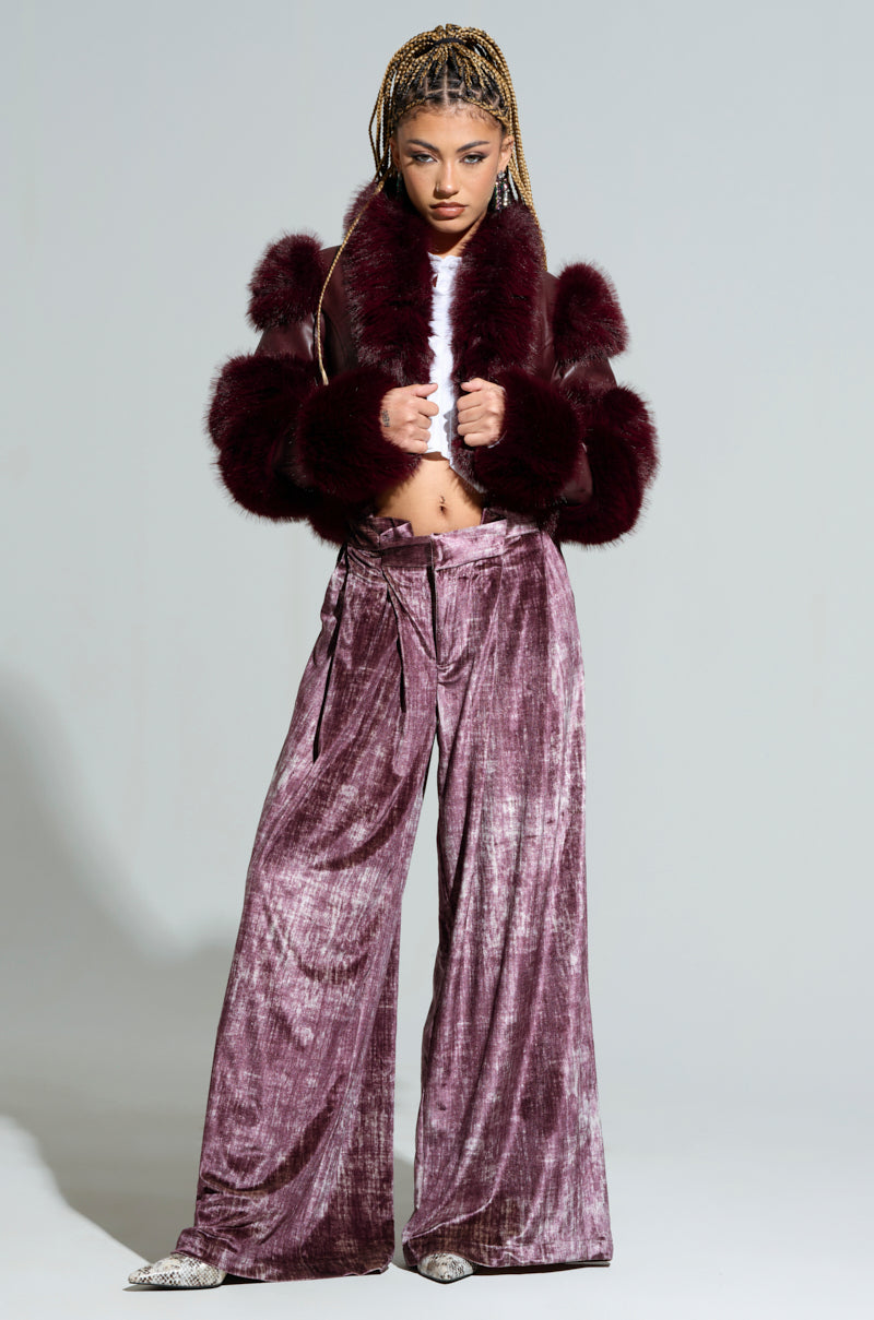 A person with braided hair poses confidently in the LOVERBOY Faux Fur Moto Jacket in burgundy, paired with velvet purple wide-leg pants. They stand against a plain background, clutching the jacket collar with both hands.