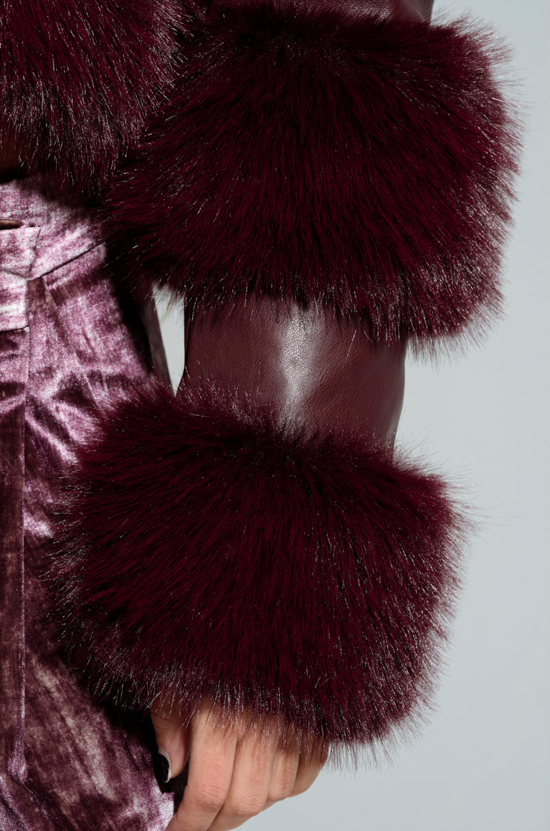 A close-up of a person's arm displays the luxurious sleeve of the LOVERBOY FAUX FUR MOTO JACKET in burgundy, paired with a soft velvet garment in matching hues. The ensemble showcases rich textures and deep colors, emphasizing a fashion-forward and opulent style with structured faux leather elements.