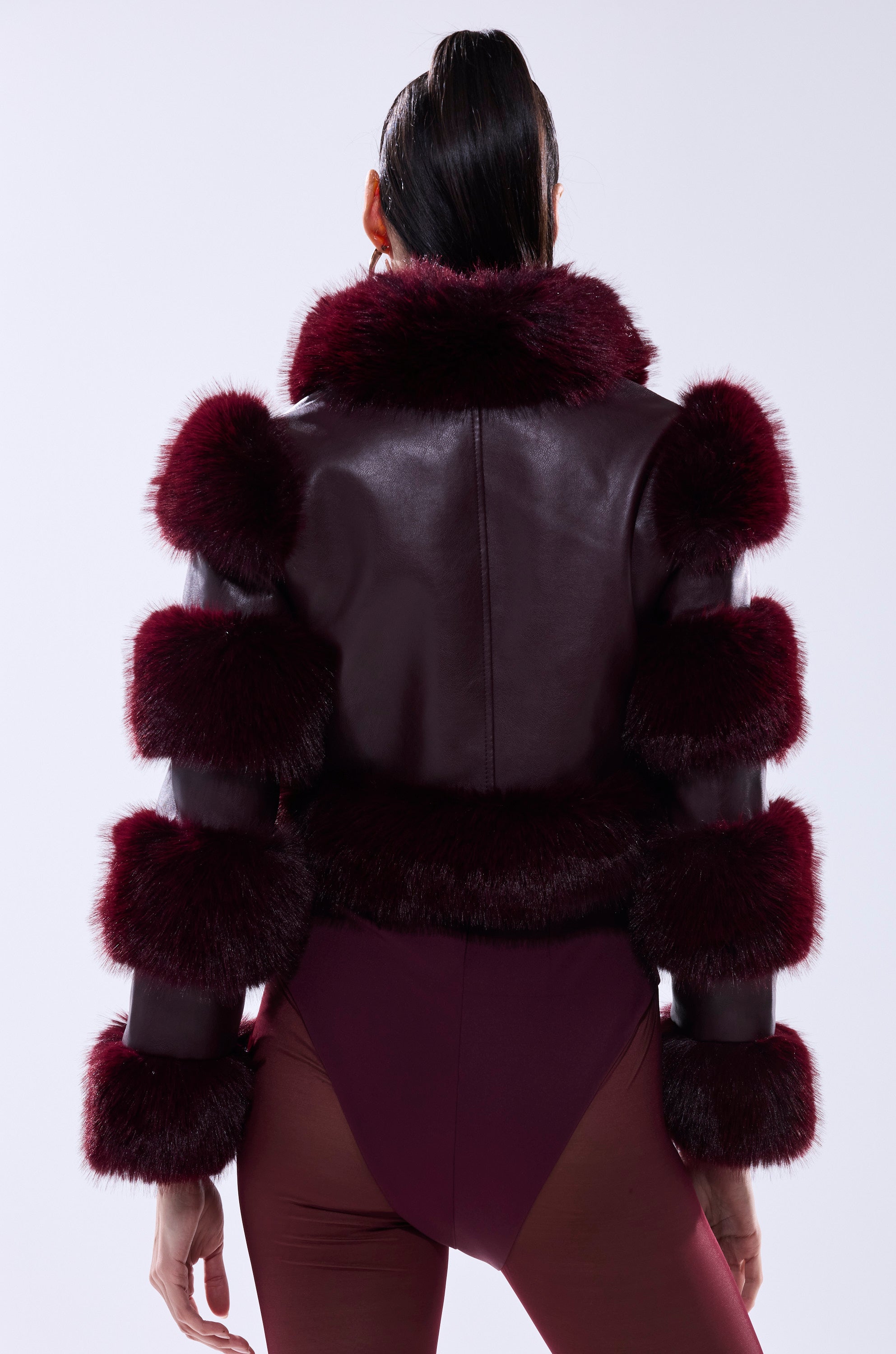 A person faces away from the camera, highlighting the LOVERBOY FAUX FUR MOTO JACKET IN BURGUNDY, featuring bold, fluffy dark red fur accents on the collar, sleeves, and hem, paired with matching burgundy bottoms.