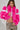 A person showcases the LOVERBOY Faux Fur Moto Jacket in pink, featuring metallic accents, paired with a cropped top and leopard print pants. They wear long braided hair, hoop earrings, and flaunt a hand tattoo while standing against a plain background.