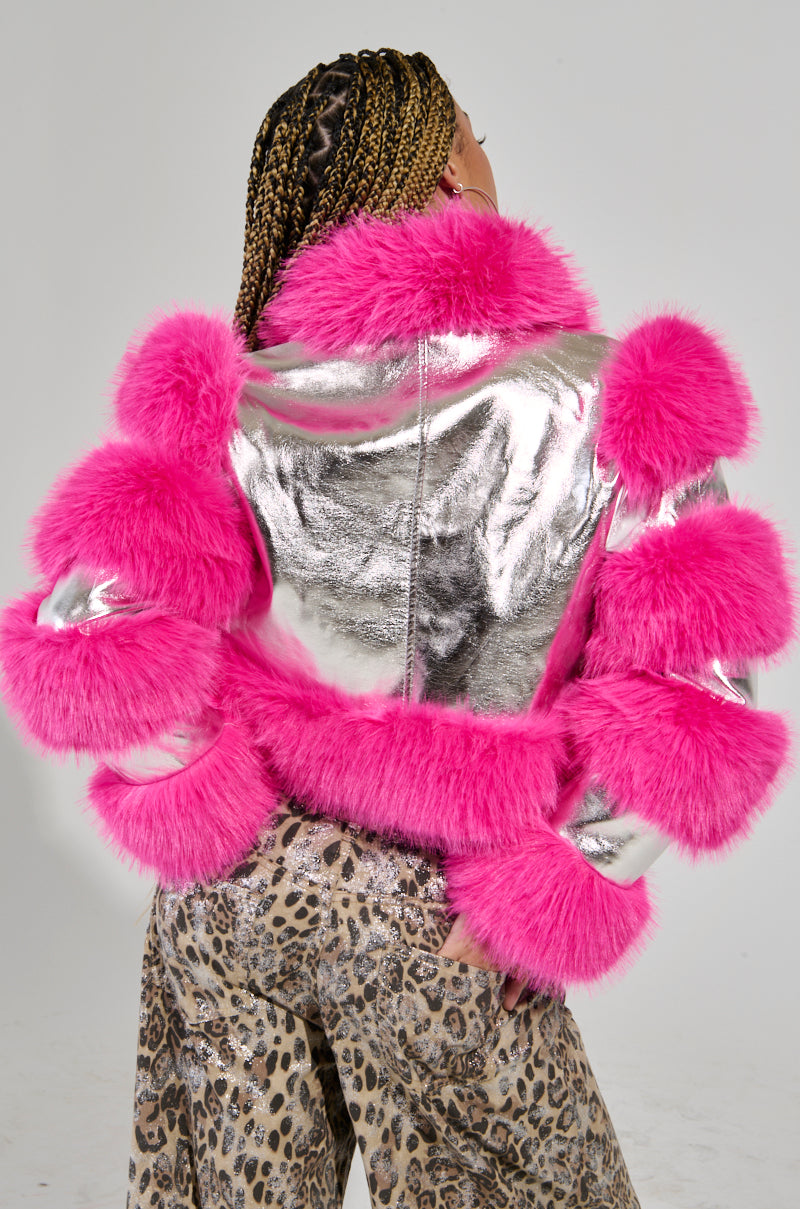 A person with braided hair wears the Loverboy Faux Fur Moto Jacket in Pink, featuring shiny silver fabric with striking pink accents on the shoulders and sleeves. Their stylish ensemble is completed with leopard print pants, set against a plain light gray background.