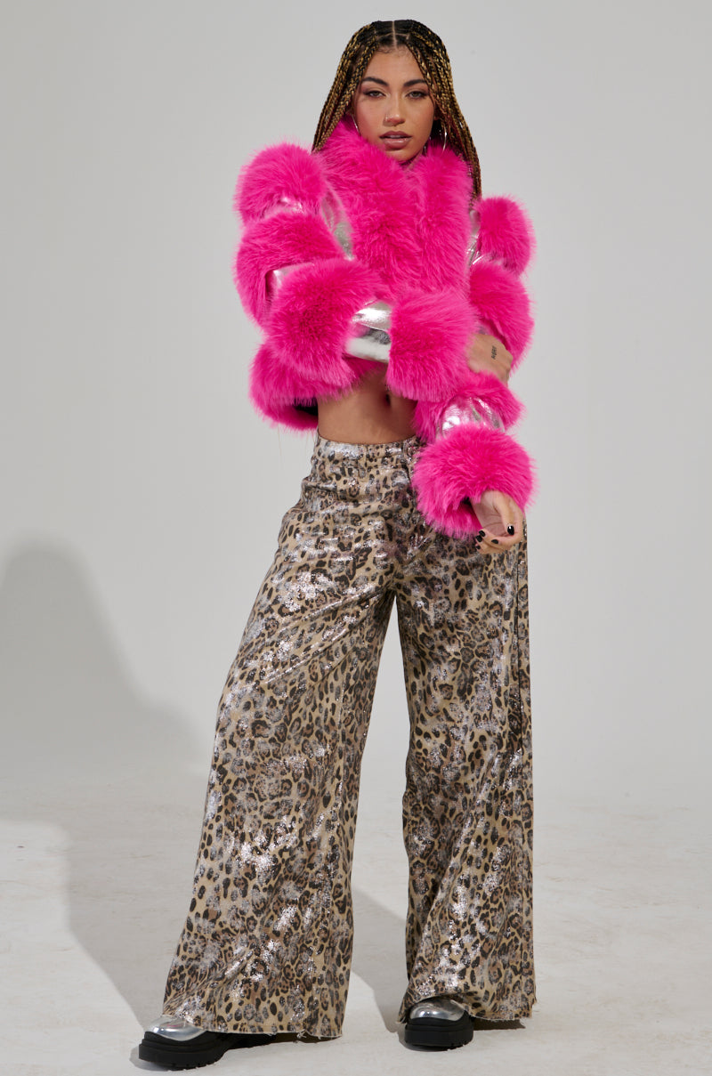 A person with braided hair poses confidently against a plain background, wearing the vibrant Loverboy Faux Fur Moto Jacket in pink, complemented by leopard print wide-leg pants and black platform shoes.