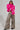 A person with braided hair poses confidently against a plain background, wearing the vibrant Loverboy Faux Fur Moto Jacket in pink, complemented by leopard print wide-leg pants and black platform shoes.