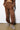 A person standing on a concrete floor wears brown sweatpants with their hands in the pockets and a matching cropped sweatshirt. Their feet are stylishly clad in the AZALEA WANG LOVING LEOPARD SANDAL, featuring a cozy leopard print design and cushioned insoles, against a backdrop of a plain white wall.