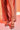 A person stands on a white floor under warm, reddish-orange lighting, wearing a light pink dress and AKIRA LOWRI BROWN SNAKE SPIRAL STILETTO SANDALS. Only their legs and feet are visible.