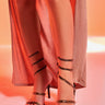 A person stands on a white floor under warm, reddish-orange lighting, wearing a light pink dress and AKIRA LOWRI BROWN SNAKE SPIRAL STILETTO SANDALS. Only their legs and feet are visible.
