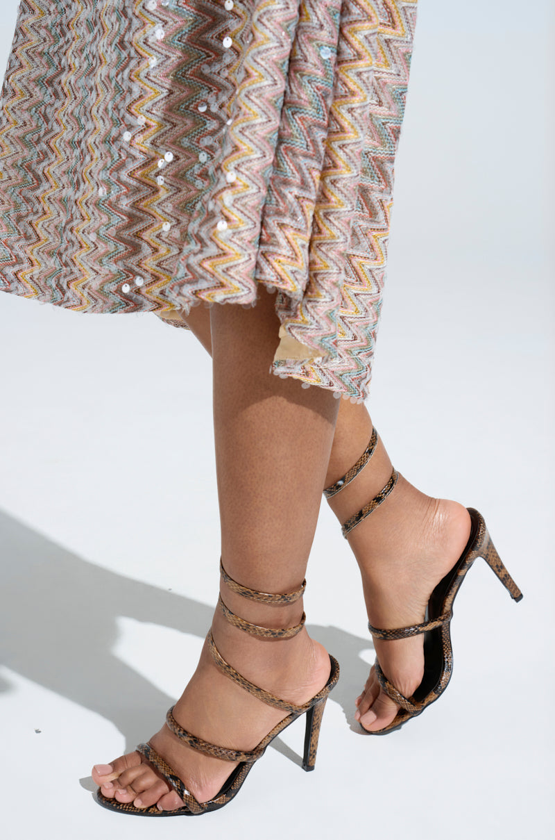 Close-up of a person wearing AKIRA LOWRI BROWN SNAKE SPIRAL STILETTO SANDALS with vegan leather in snakeskin print and wraparound ankle straps, paired with a colorful zigzag-patterned skirt against a plain light background.