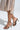 Close-up of a person wearing AKIRA LOWRI BROWN SNAKE SPIRAL STILETTO SANDALS with vegan leather in snakeskin print and wraparound ankle straps, paired with a colorful zigzag-patterned skirt against a plain light background.