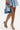A woman in a textured blue mini dress wears AZALEA WANG LOWRI DENIM SPARKLE COIL WRAP UP SANDALS and holds a small patchwork denim handbag against a plain white background.