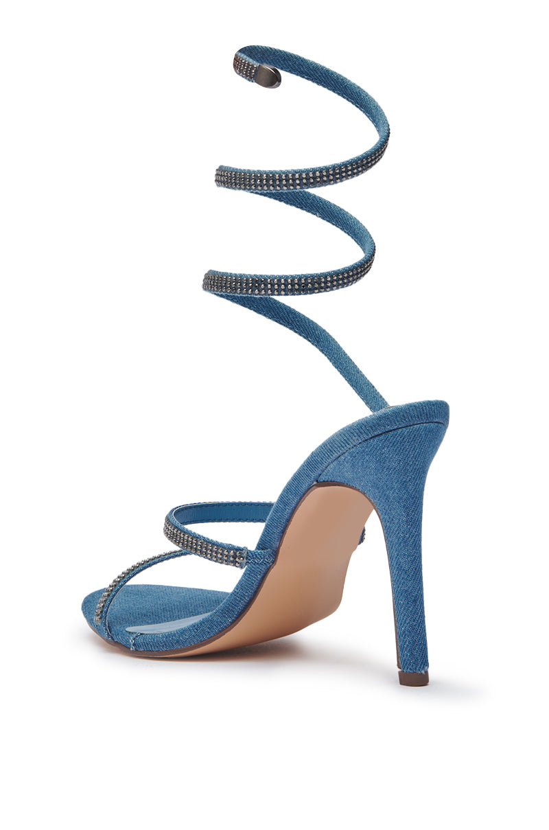 The AZALEA WANG LOWRI DENIM SPARKLE COIL WRAP UP SANDAL features a blue denim design, stiletto heel, rhinestone-embellished spiral ankle strap, and two rhinestone-studded front straps. Denim heel and sole pop against a white background. 5