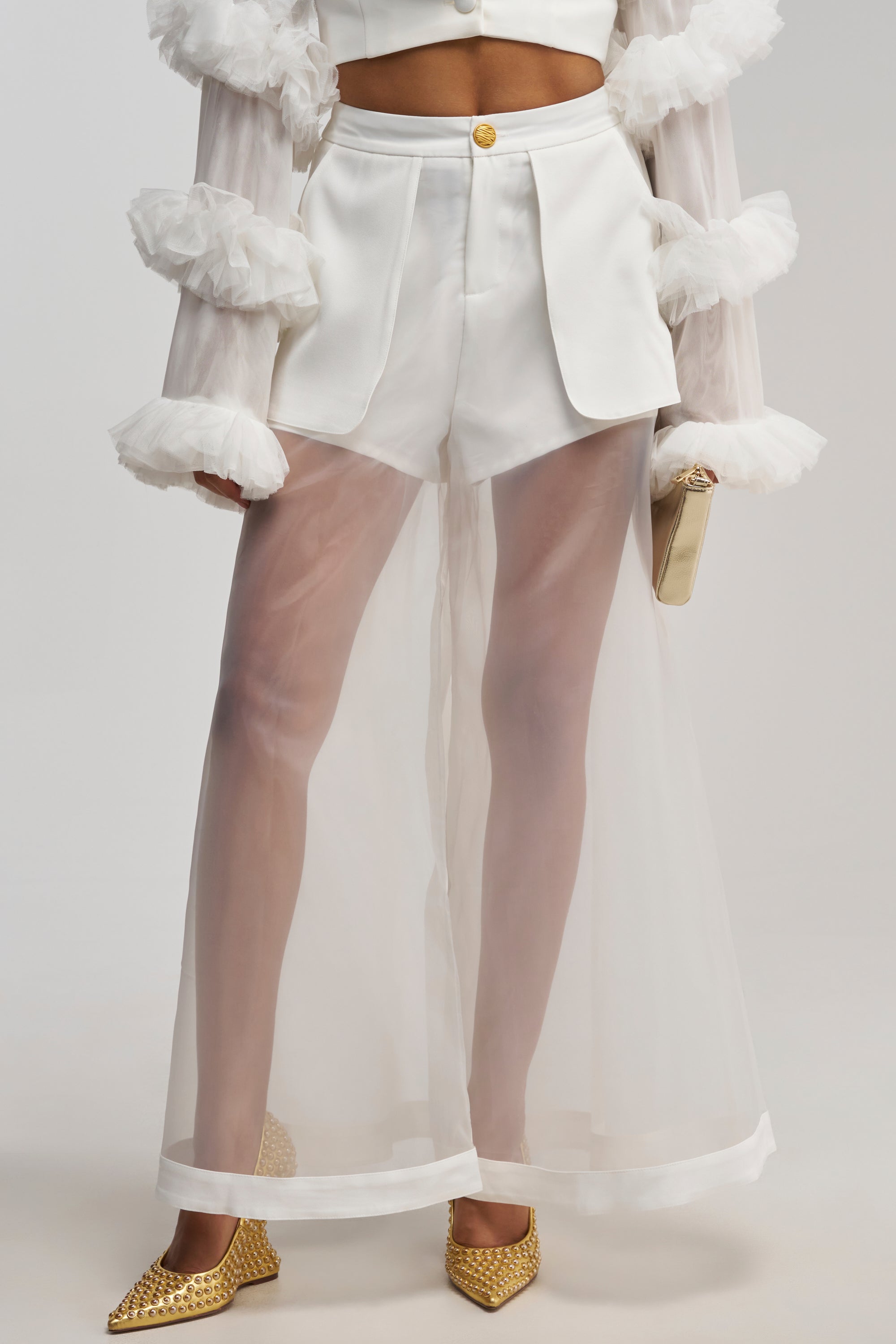 A woman wears the DRESSED TO IMPRESS TROUSER, styled in a white high-waisted sheer mesh wide-leg fit, paired with a white crop top with ruffled sleeves. She holds a clutch and completes the look with gold pointed-toe shoes.
