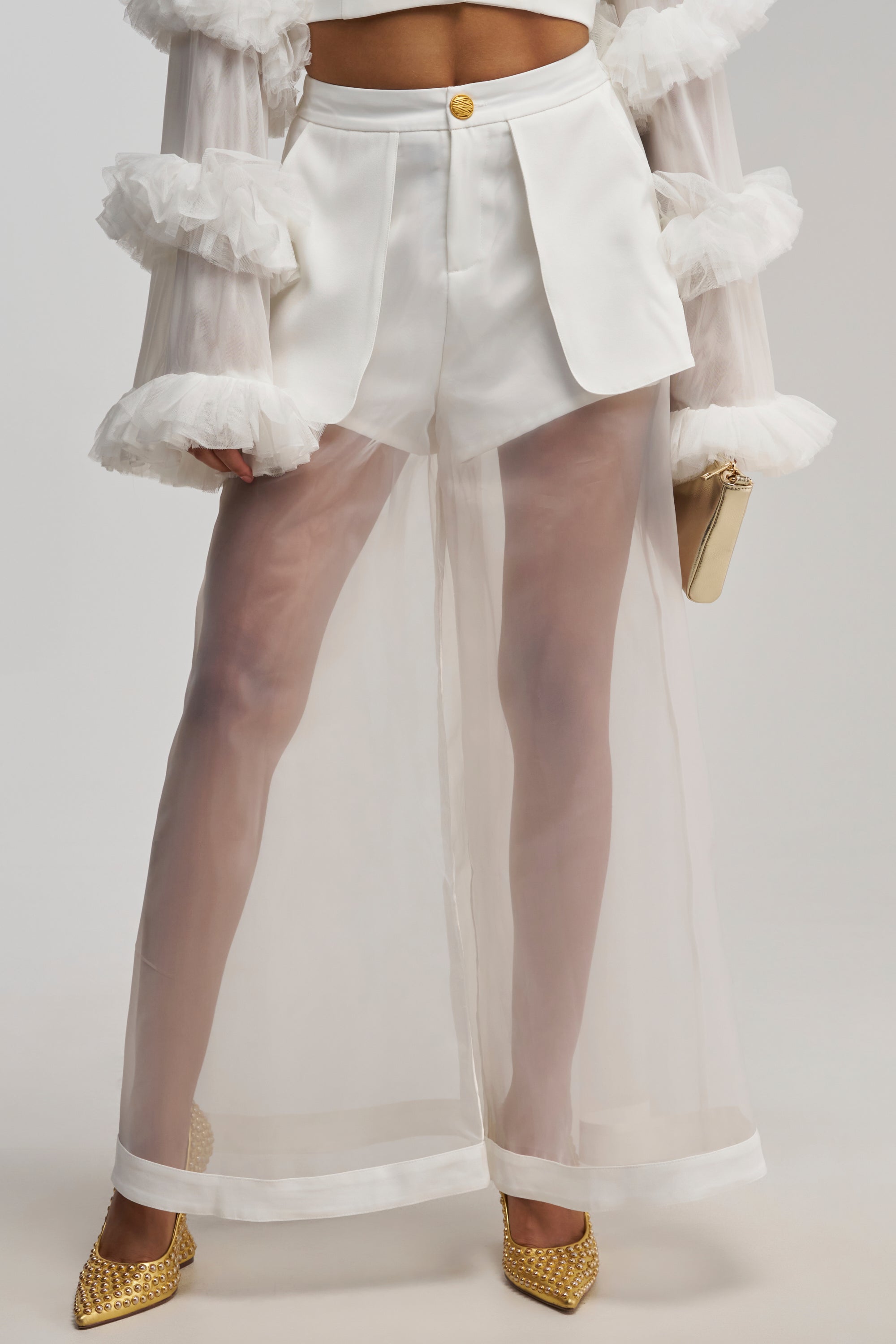 Wearing the DRESSED TO IMPRESS TROUSER, which features white tailored shorts with a sheer mesh, floor-length wide-leg overlay, paired with a ruffled sheer white top, gold pointed heels, and a small beige clutch.