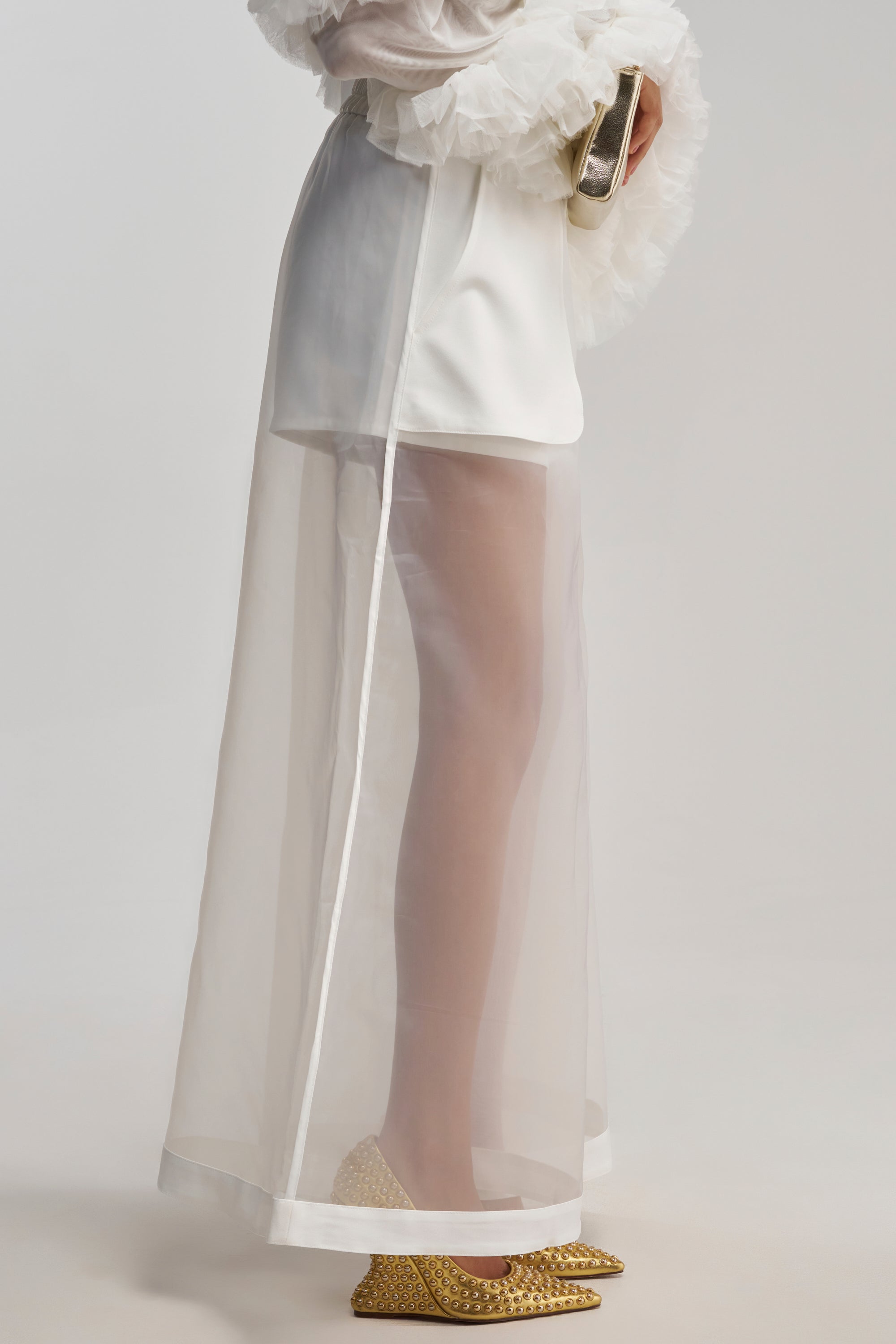 Wearing the DRESSED TO IMPRESS TROUSER, styled with a white blouse featuring ruffled sleeves, a small silver clutch, and gold pointed-toe heels.