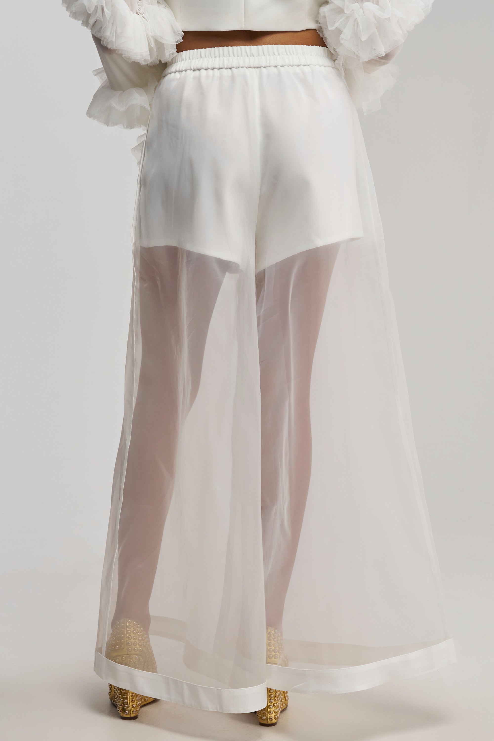 Wearing the DRESSED TO IMPRESS TROUSER in a sheer, wide-leg fit over white shorts and gold shoes, this back view showcases translucent, flowy trousers that reveal the shorts underneath. The look is completed with a top featuring ruffled sleeves.