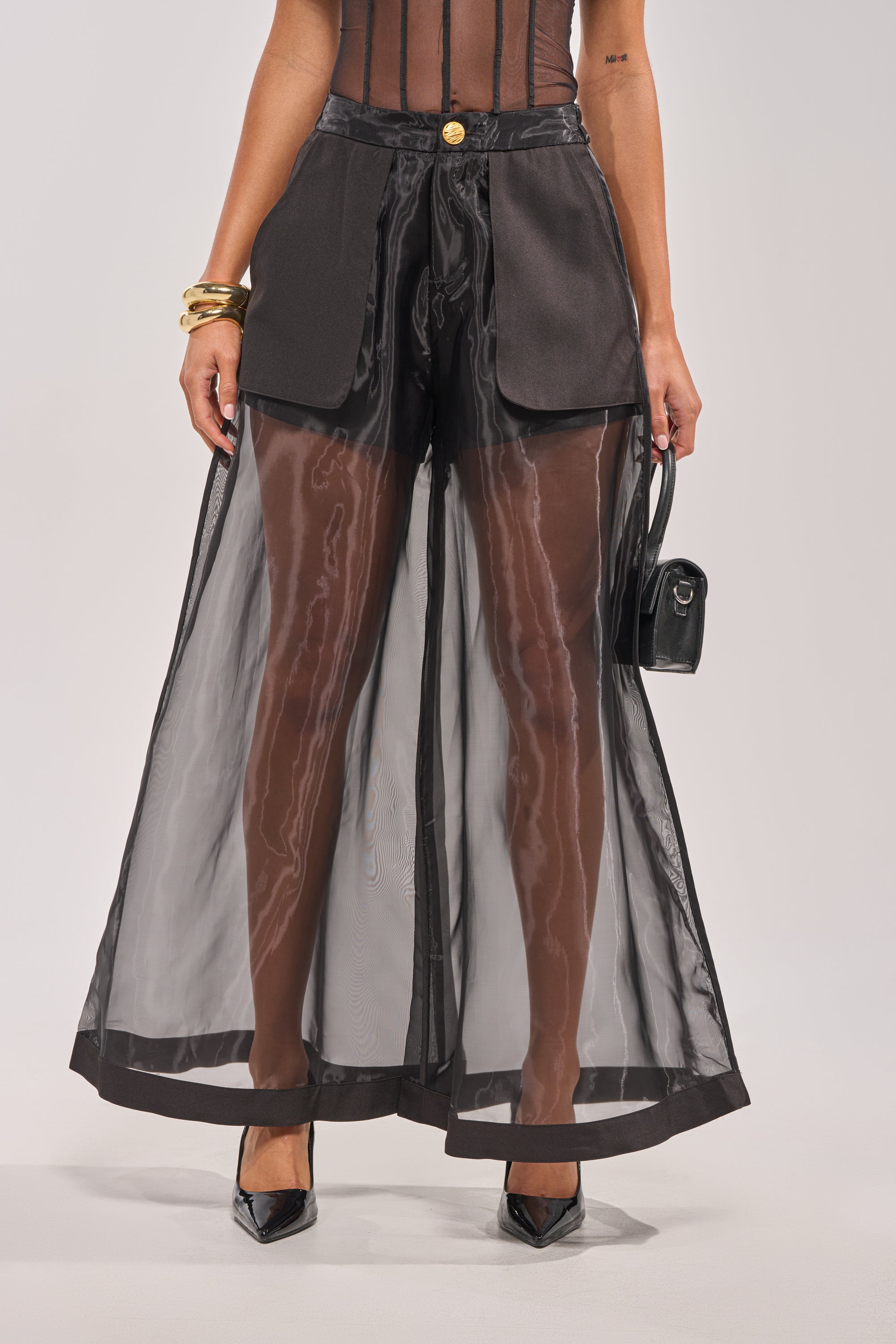 A woman styles the FANCY LIKE ME TROUSER—black mesh wide-leg pants with a high-rise waistband and solid black shorts underneath—with a black mesh top, black heels, gold bracelets, and a small black handbag. 2