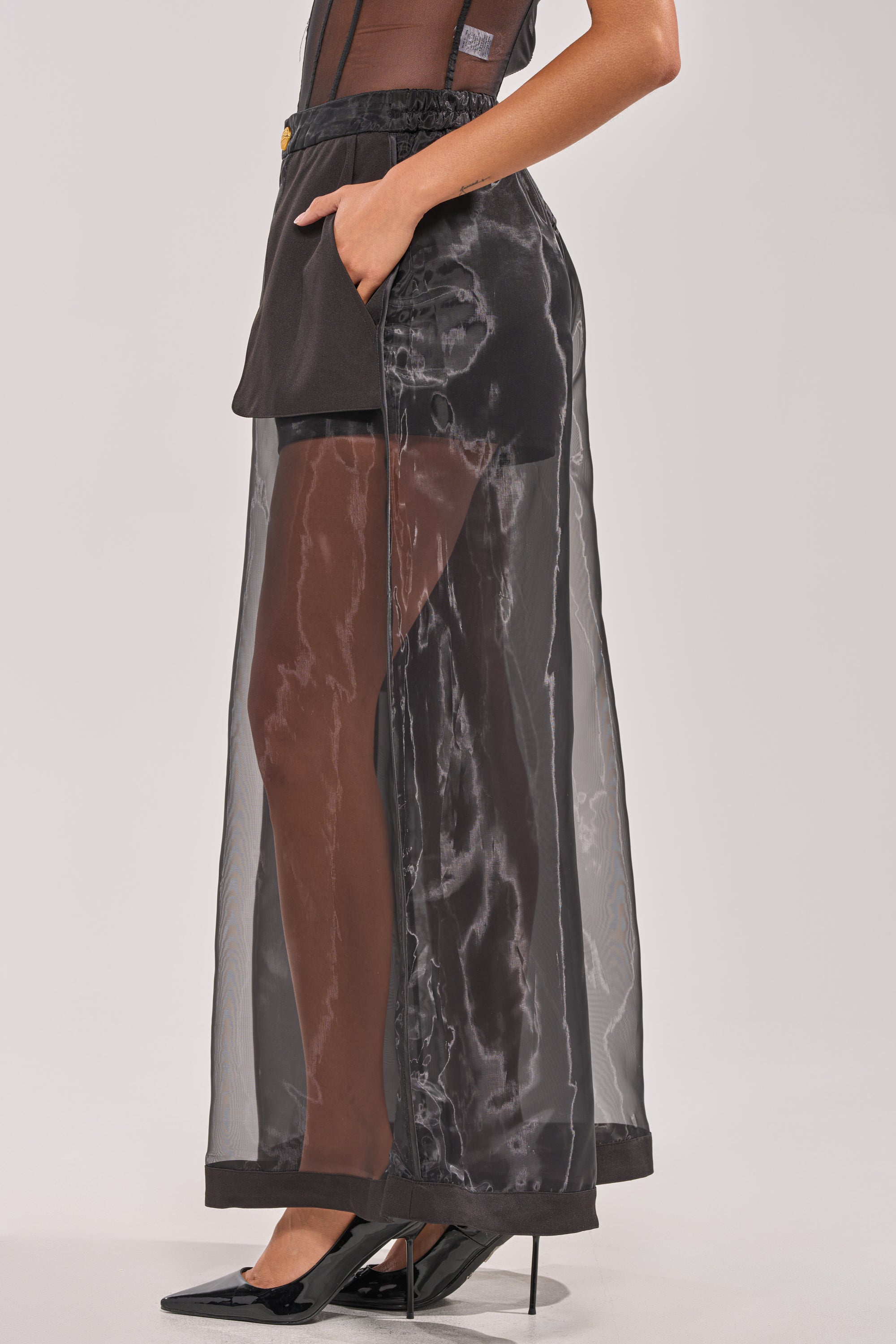 A person wears the FANCY LIKE ME TROUSER, featuring black mesh wide-leg pants over black high-waisted shorts, with a sleeveless top and black pointed-toe heels. One hand is in the pocket. The background is plain light gray. 3