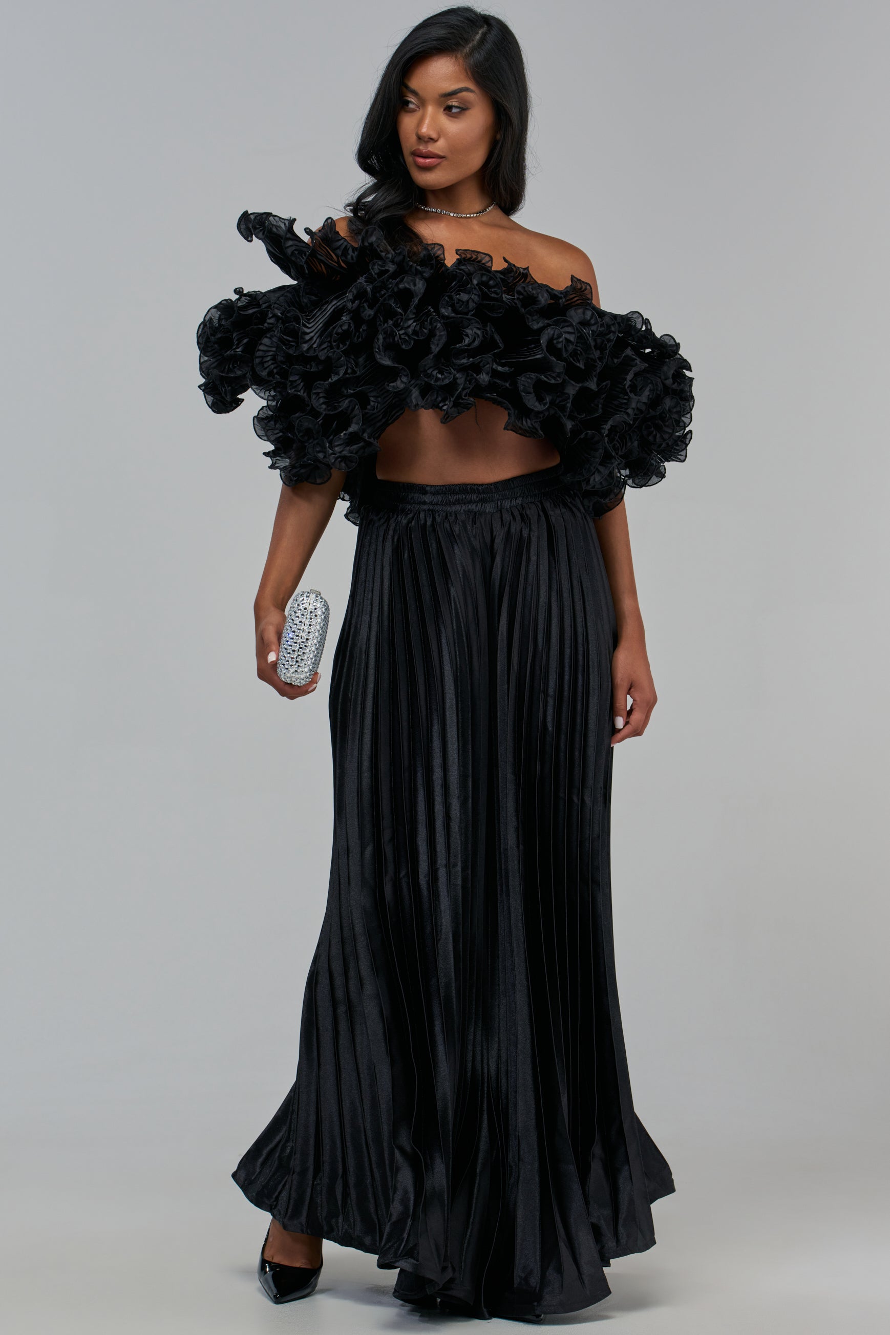 A person wears an off-the-shoulder black top with a large ruffled neckline and black satin BIG CITY RUFFLE PANT, holding a small silver embellished clutch.