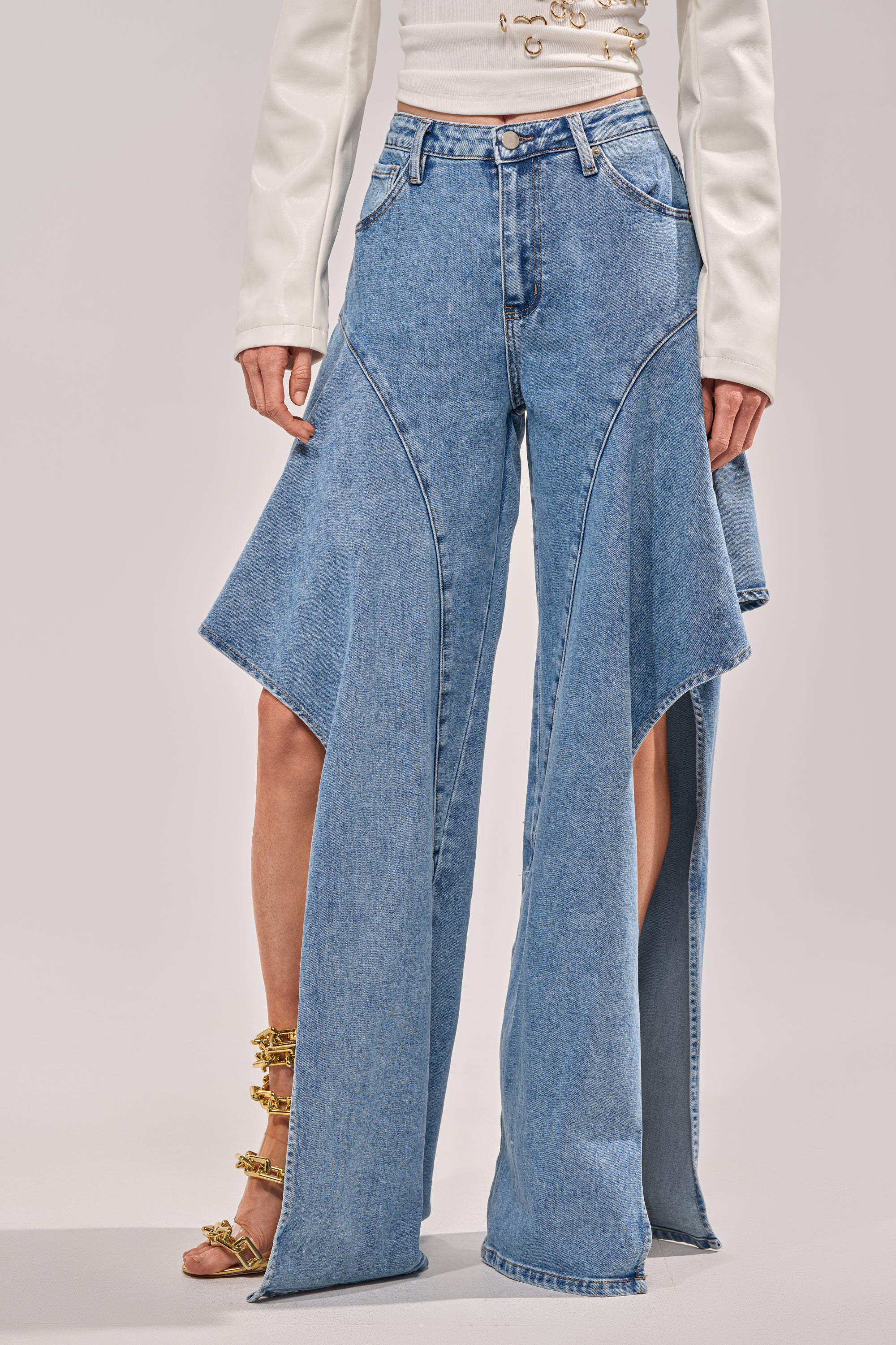 A person wears the CHECK IT OUT RELAXED DENIM, featuring high-rise blue jeans with wide flared panels, styled with a white long-sleeve top and gold strappy heels. 1