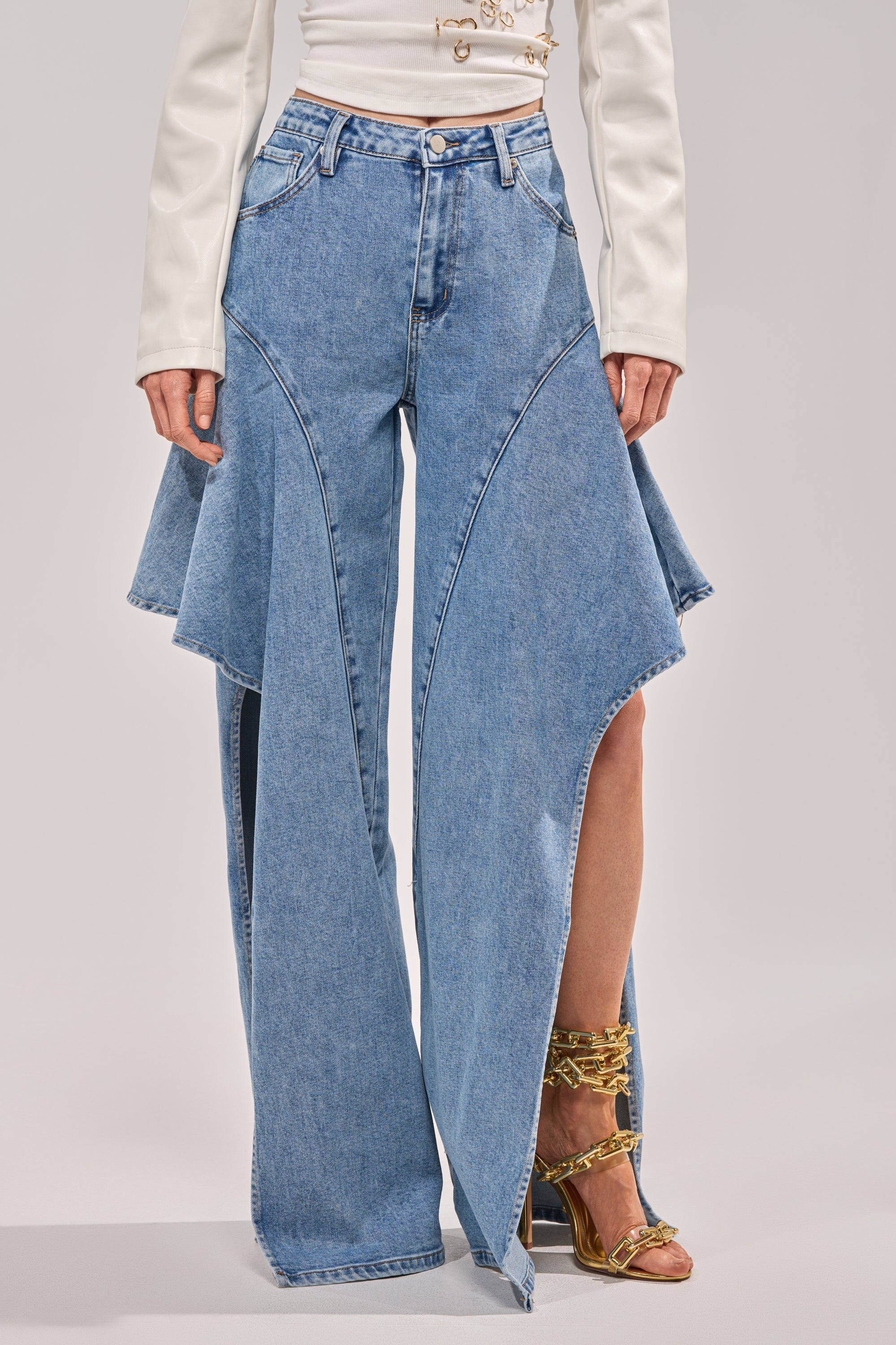 A person wearing CHECK IT OUT RELAXED DENIM, high-rise wide-leg jeans with dramatic side panels and high slits, paired with gold strappy heels and a white long-sleeve top. 2