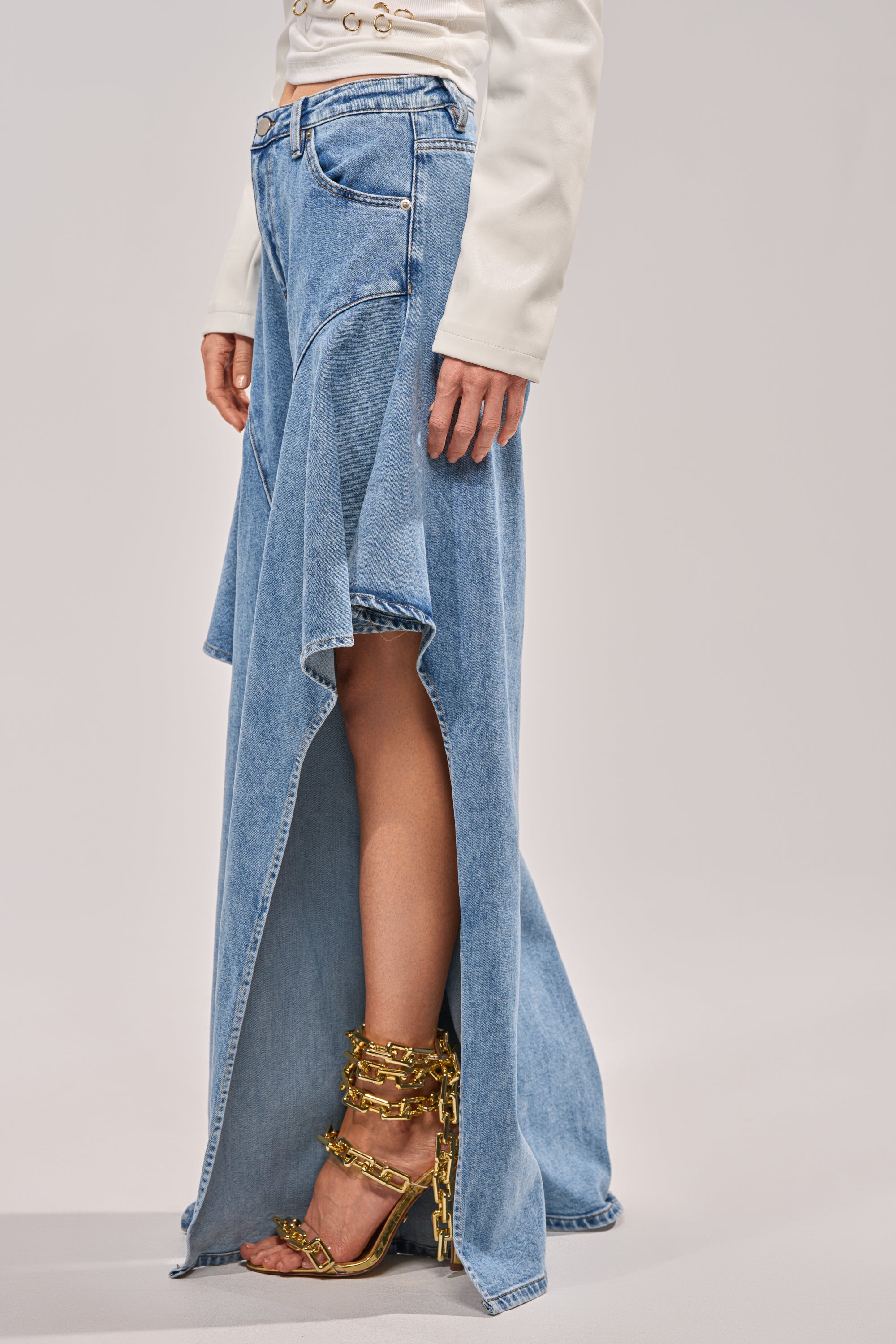 Someone wears the CHECK IT OUT RELAXED DENIM long asymmetrical medium wash skirt with a high front slit, paired with a white long-sleeve top and black sandals adorned with gold chain details. 3