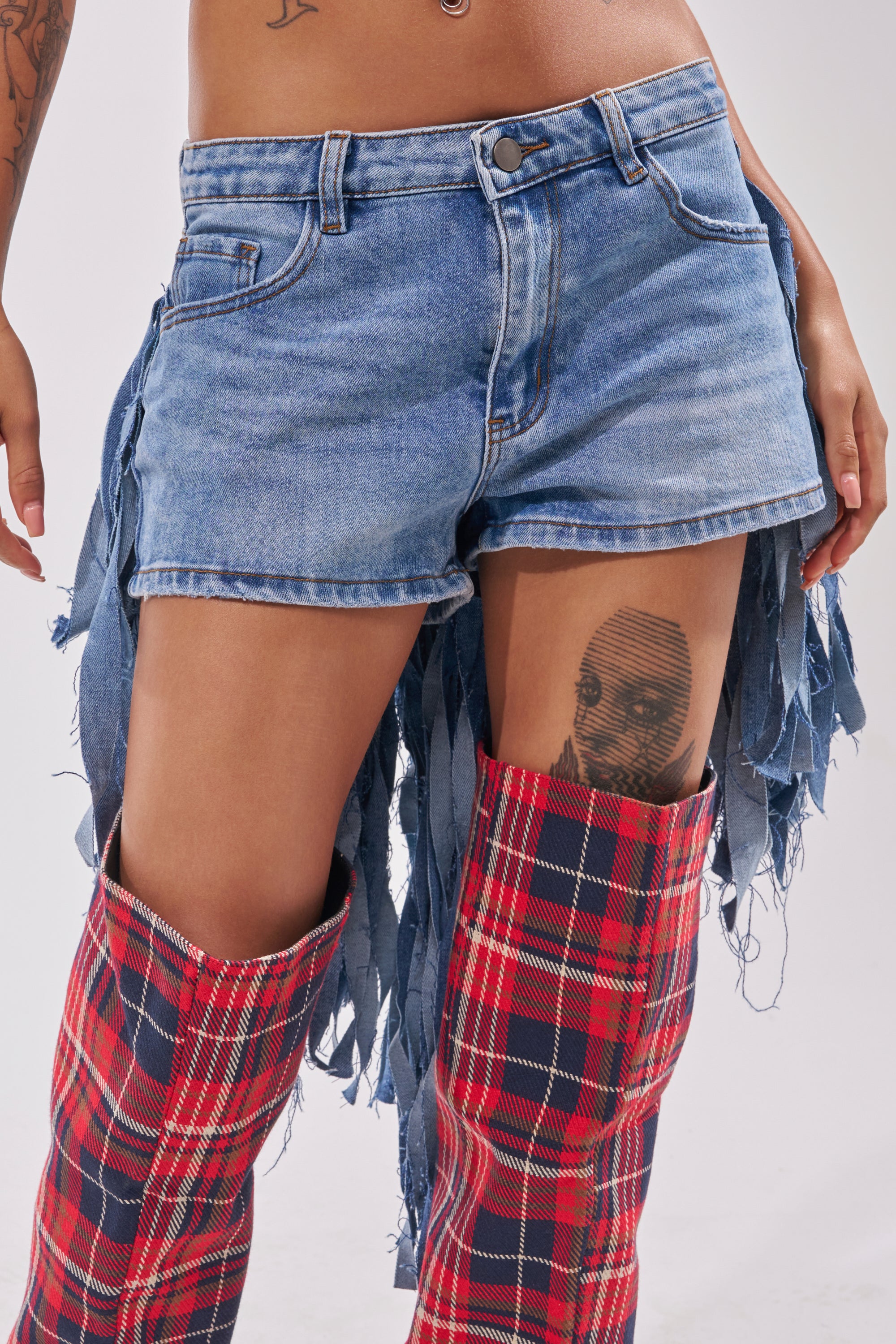 Wearing TURN AROUND DISTRESSED DENIM SHORTS with frayed sides, the person pairs them with red plaid thigh-high boots. A face tattoo is visible on their upper thigh against a plain, light-colored background. 0