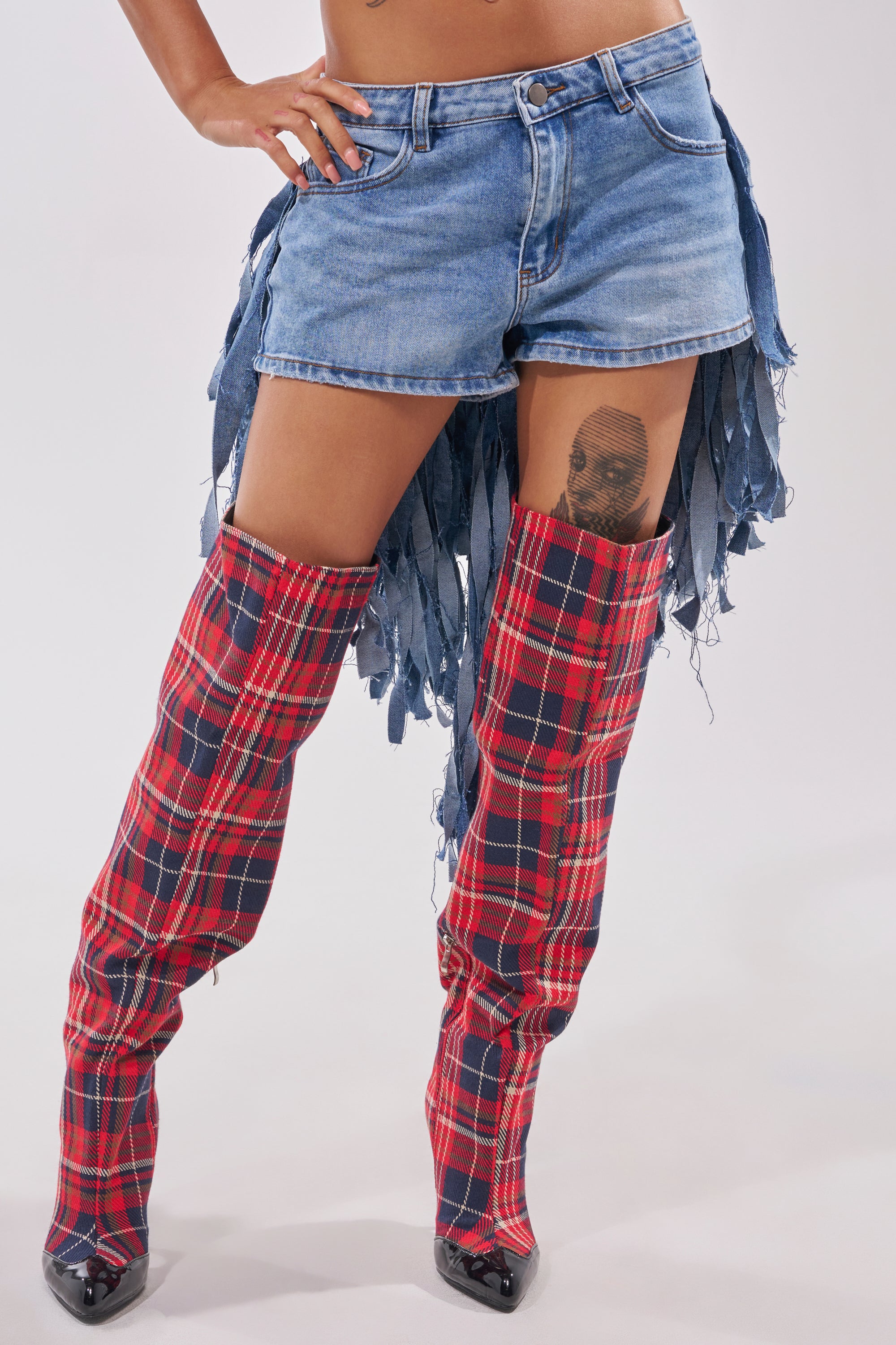Wearing the TURN AROUND DISTRESSED DENIM SHORT with frayed strips and red plaid over-the-knee pointed-toe boots, a person reveals a tattoo on their thigh. 1