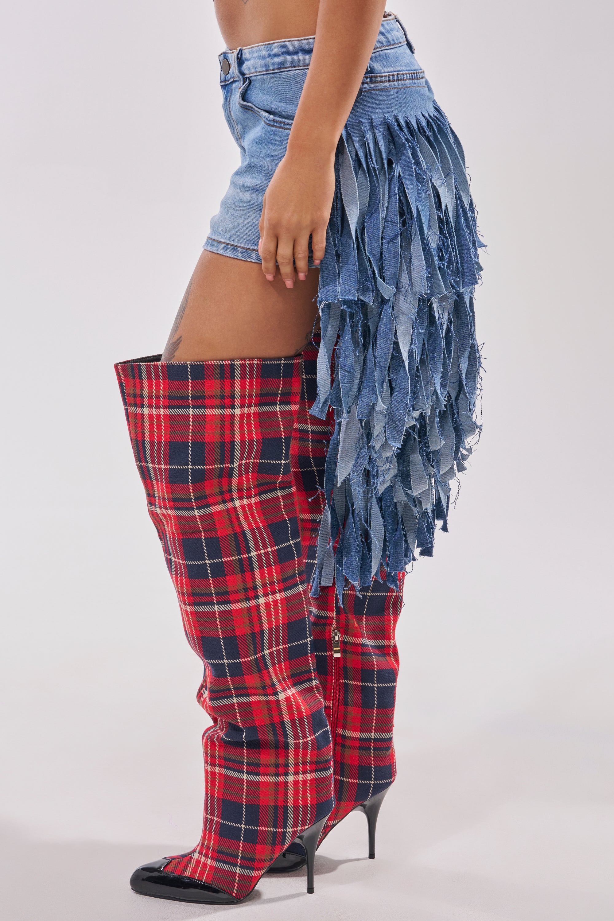 Someone wearing the TURN AROUND DISTRESSED DENIM SHORT, featuring long frayed fringe on one side, paired with thigh-high red plaid stiletto boots—a bold festival look seen from the side against a plain background. 2