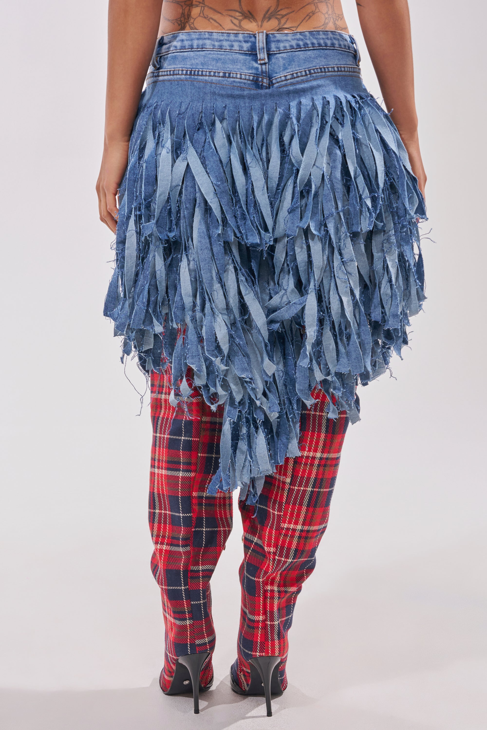 A person stands facing away, wearing black heels, red plaid pants, and the TURN AROUND DISTRESSED DENIM SHORT with long, shredded fringe. Their lower back tattoo is partly visible above the western-inspired shorts. 3