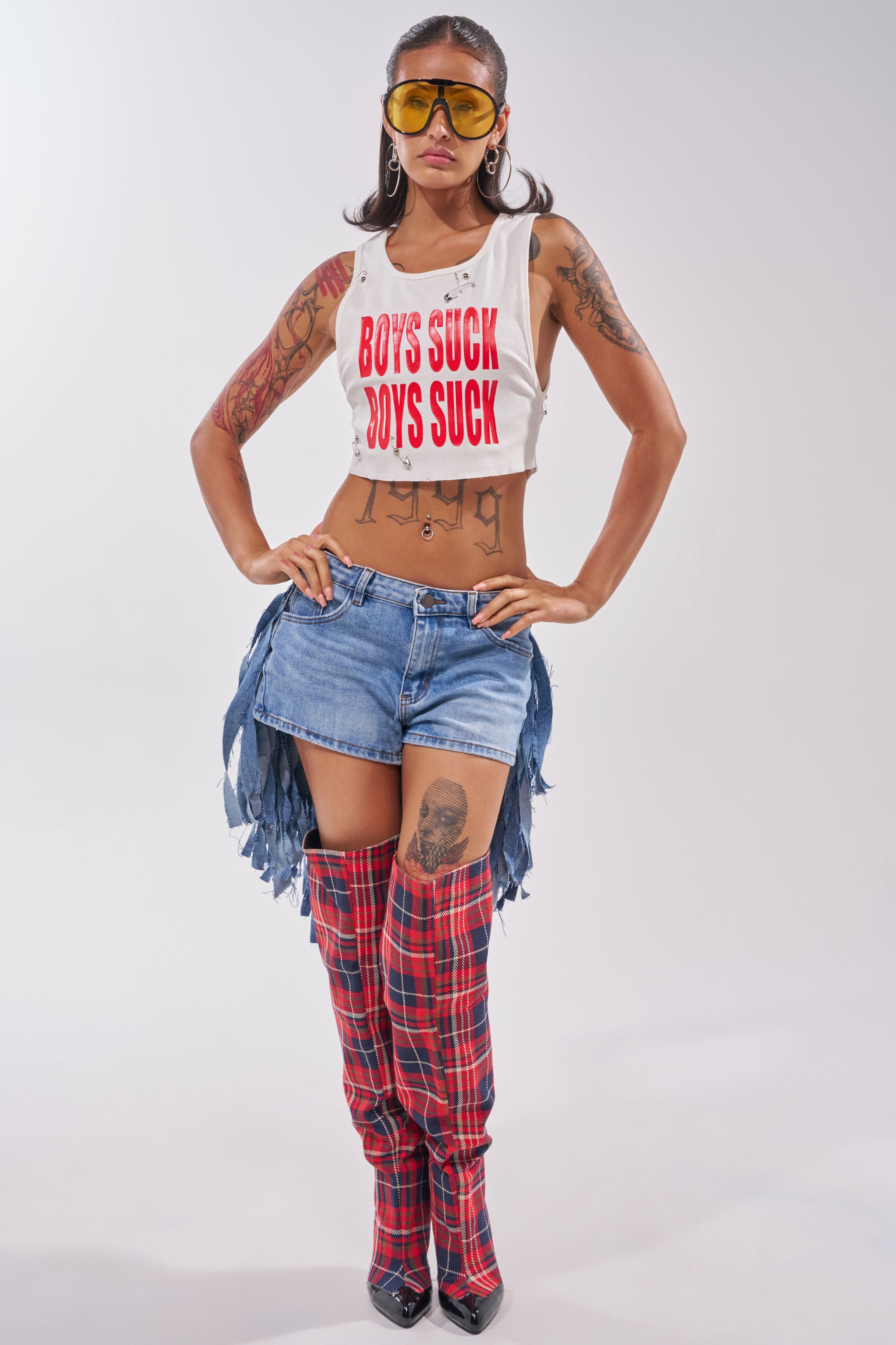 A woman with tattoos poses confidently in yellow sunglasses, a "BOYS SUCK" crop top, TURN AROUND DISTRESSED DENIM SHORTS, and red plaid thigh-high boots. 4