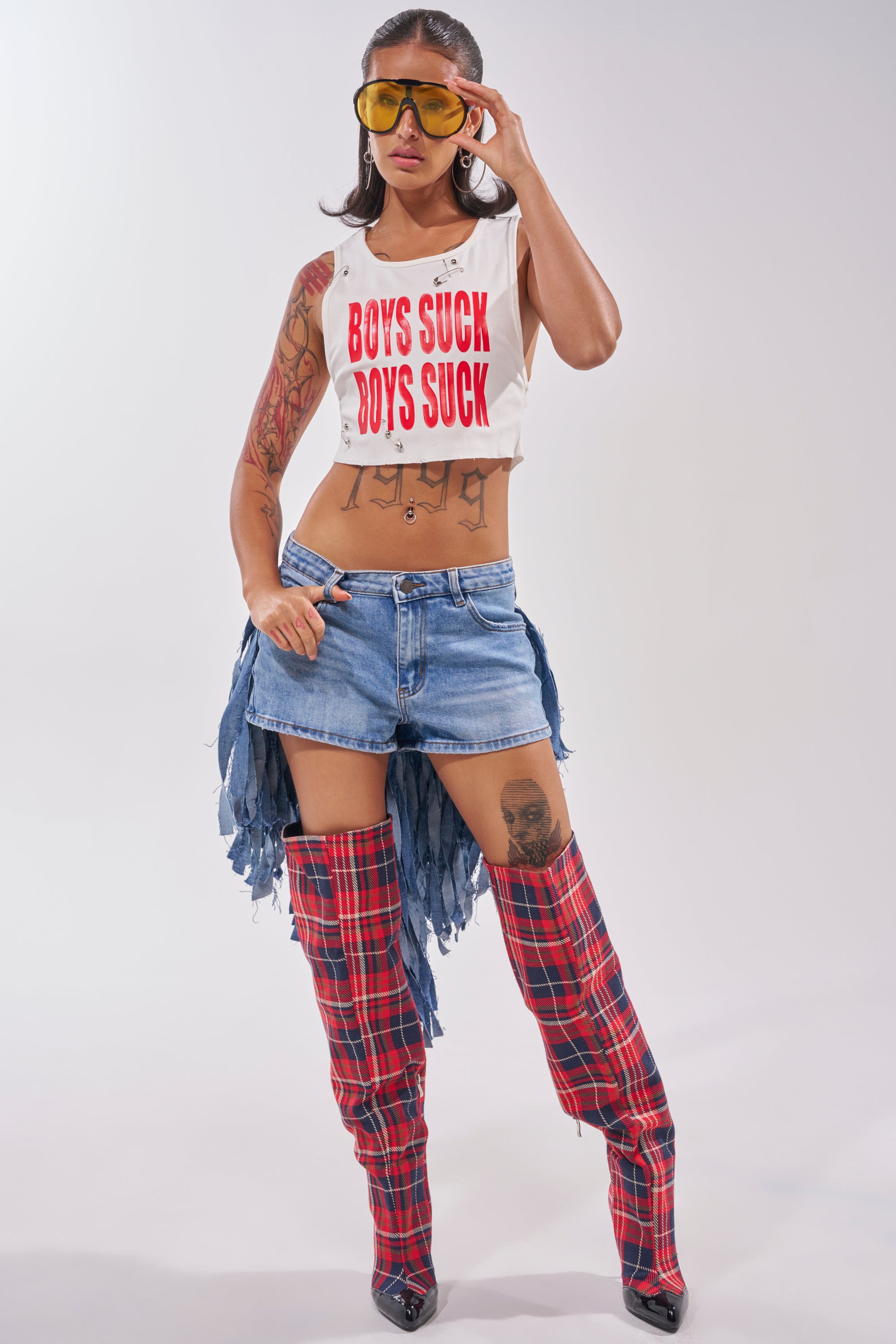 A woman models yellow sunglasses, a "BOYS SUCK" crop top, red plaid thigh-high boots, and the TURN AROUND DISTRESSED DENIM SHORT. She has torso and leg tattoos and casually tucks one hand into her pocket. 6