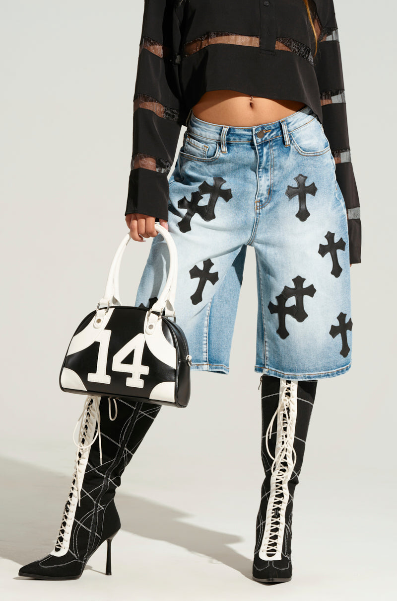 A person dressed in trendy fashion showcases the "CROSS YOU OFF BERMUDA DENIM SHORT," adorned with black cross designs on a light blue fabric. They pair these chic high-rise shorts with knee-high black lace-up boots and a long-sleeve black mesh crop top, accessorizing with a stylish black and white handbag featuring the number 14 in bold white. 2