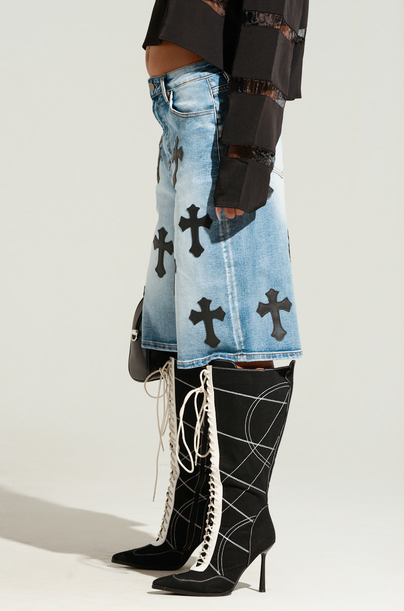 A person wearing a black long-sleeve top, light blue CROSS YOU OFF BERMUDA DENIM SHORT adorned with black cross and faux leather patches, and high-heeled, black knee-high boots with white lace-up detailing. The high-rise design of the outfit is contemporary and stylish, blending gothic and modern elements. 3