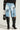 A person is wearing the CROSS YOU OFF Bermuda denim shorts in light blue, featuring black faux leather patches. They complement their outfit with black knee-high boots that have white lace-up designs and carry a stylish black and white handbag. The background is plain.