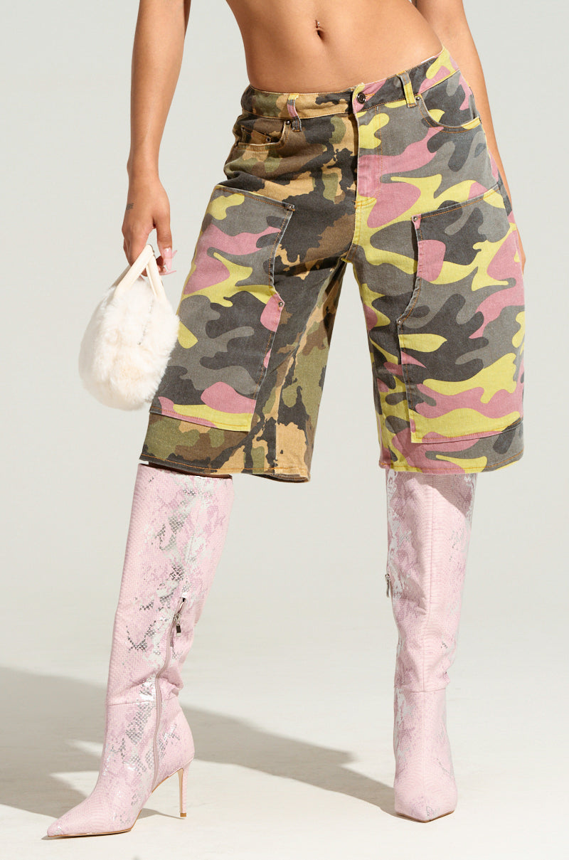 Sporting the GO TIME CAMO SHORT in a patchwork of green, yellow, and pink, paired with light pink knee-high textured boots, this person holds a small, fluffy white handbag as they stand against a plain background. Only their lower body is visible.