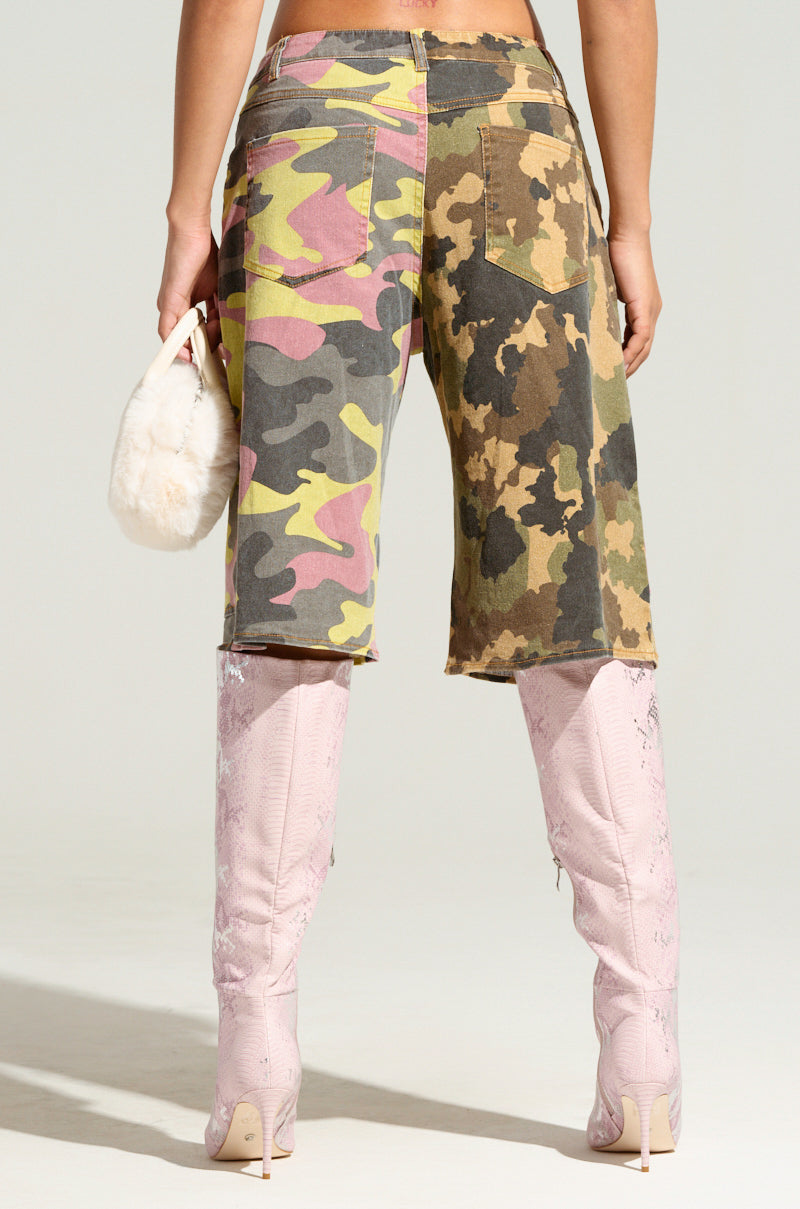 A person stands facing away, embracing utility style with the GO TIME CAMO SHORTS and thigh-high pink embossed boots. They clutch a small, fluffy white handbag against the plain gray backdrop.