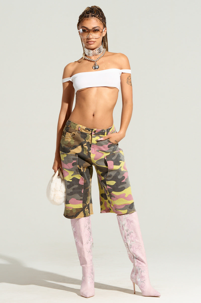 A person with braided hair stands confidently in a white off-shoulder crop top, GO TIME CAMO SHORT exuding utility style, pink knee-high boots, sunglasses, and layered necklaces. They hold a fluffy white purse against a plain background.