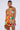 A woman is wearing the Spicy Mami Printed Romper, a colorful, sleeveless, halter-neck outfit with a cut-out detail in the midriff. The fabric features a vibrant abstract print. She holds a small woven clutch and has her hair styled in a sleek ponytail.