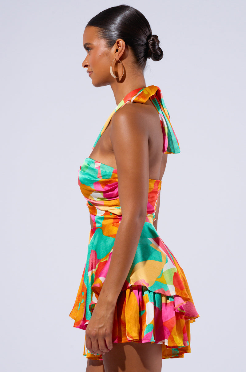 A woman stands sideways, highlighting the SPICY MAMI PRINTED ROMPER which features a colorful, multi-layered design with vibrant abstract patterns in green, pink, yellow, and orange hues. The romper boasts a sleeveless style with a halter neck tied into a knot. She accessorizes with large hoop earrings and has her hair sleekly tied back. 1