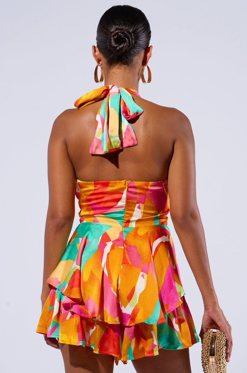 A person with their back to the camera is wearing the SPICY MAMI PRINTED ROMPER, which boasts a strapless design and a vibrant abstract pattern in orange, yellow, pink, and blue. They accessorize with large hoop earrings and hold a wicker purse in their right hand. 2