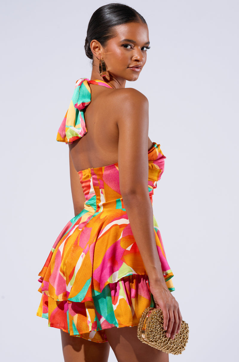 A woman wearing the SPICY MAMI PRINTED ROMPER, featuring a vibrant abstract print of pink, yellow, green, and orange, stands against a plain background. She holds a small woven clutch and looks over her shoulder towards the camera. 3