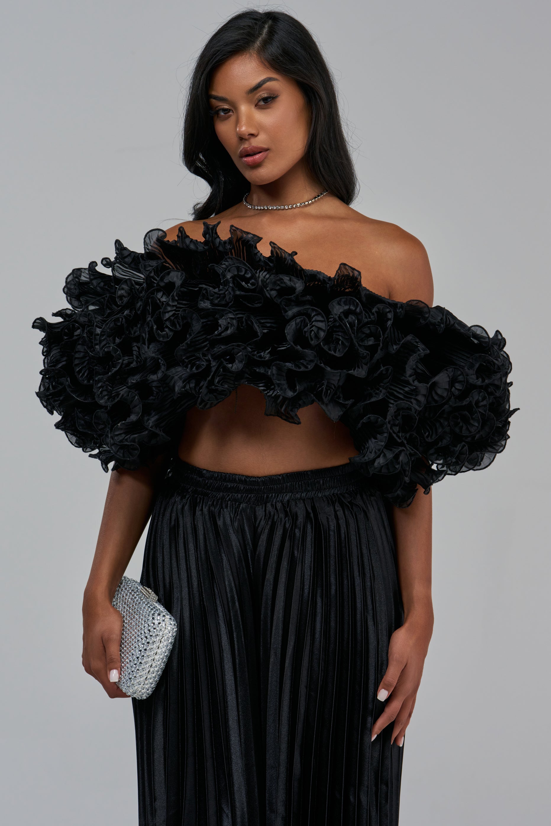 A woman wears a BIG CITY RUFFLE TOP with a sculptural black off-the-shoulder neckline, paired with a pleated black skirt and a silver clutch.