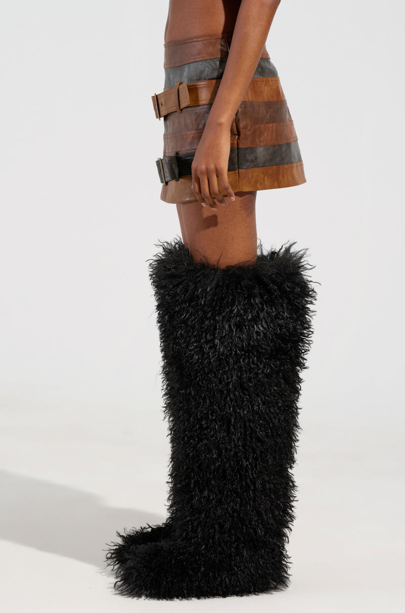 A person wearing the MESS AROUND MINI SKIRT, designed in brown faux leather with striped panel construction, pairs it with large black shaggy boots while standing against a plain background. The image captures this AKIRA Label ensemble from the side view. 1