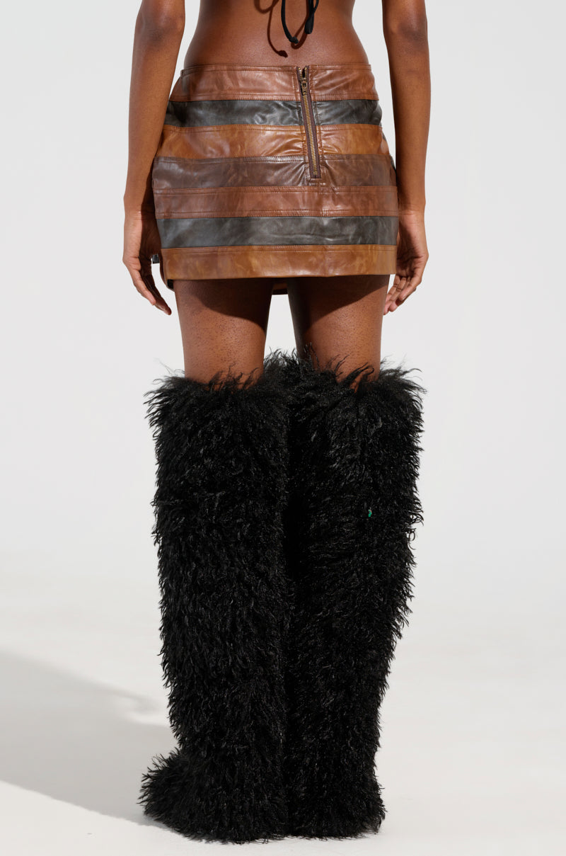 A person wearing a MESS AROUND MINI SKIRT by AKIRA Label, featuring a back zipper, stands with their back turned. They are also wearing tall, black fluffy boots that cover most of their legs. The backdrop is plain and light-colored. 2