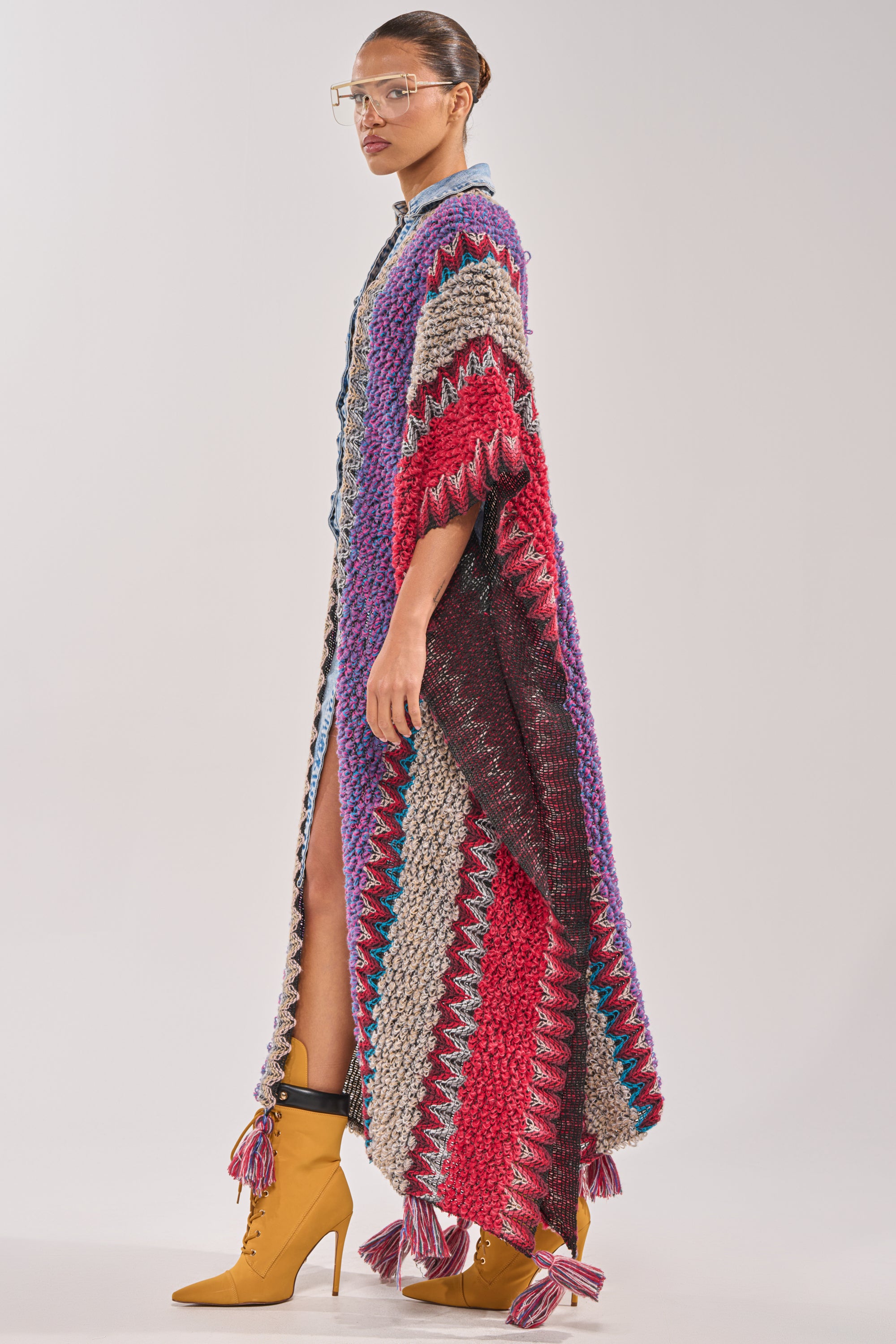 A person stands sideways wearing the JUST WHAT I AM KIMONO, a longline knit with zigzag patterns in red, purple, gray, and blue. They're paired with yellow high-heeled boots and large clear glasses. 1
