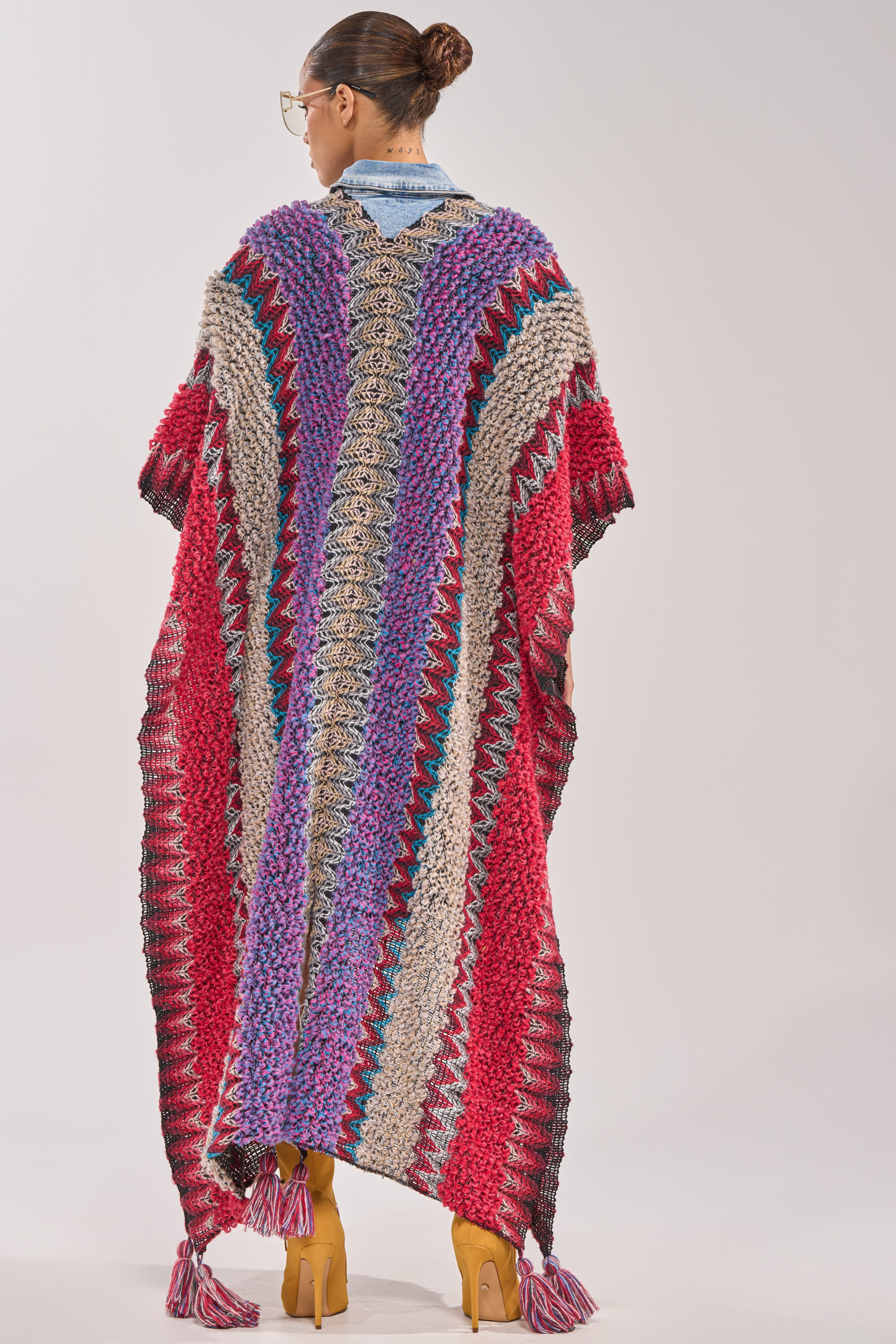 A person with a bun and glasses faces away, modeling the JUST WHAT I AM KIMONO—a colorful, zigzag-patterned festival poncho with tasseled hem in red, purple, blue, beige, and gray. 2