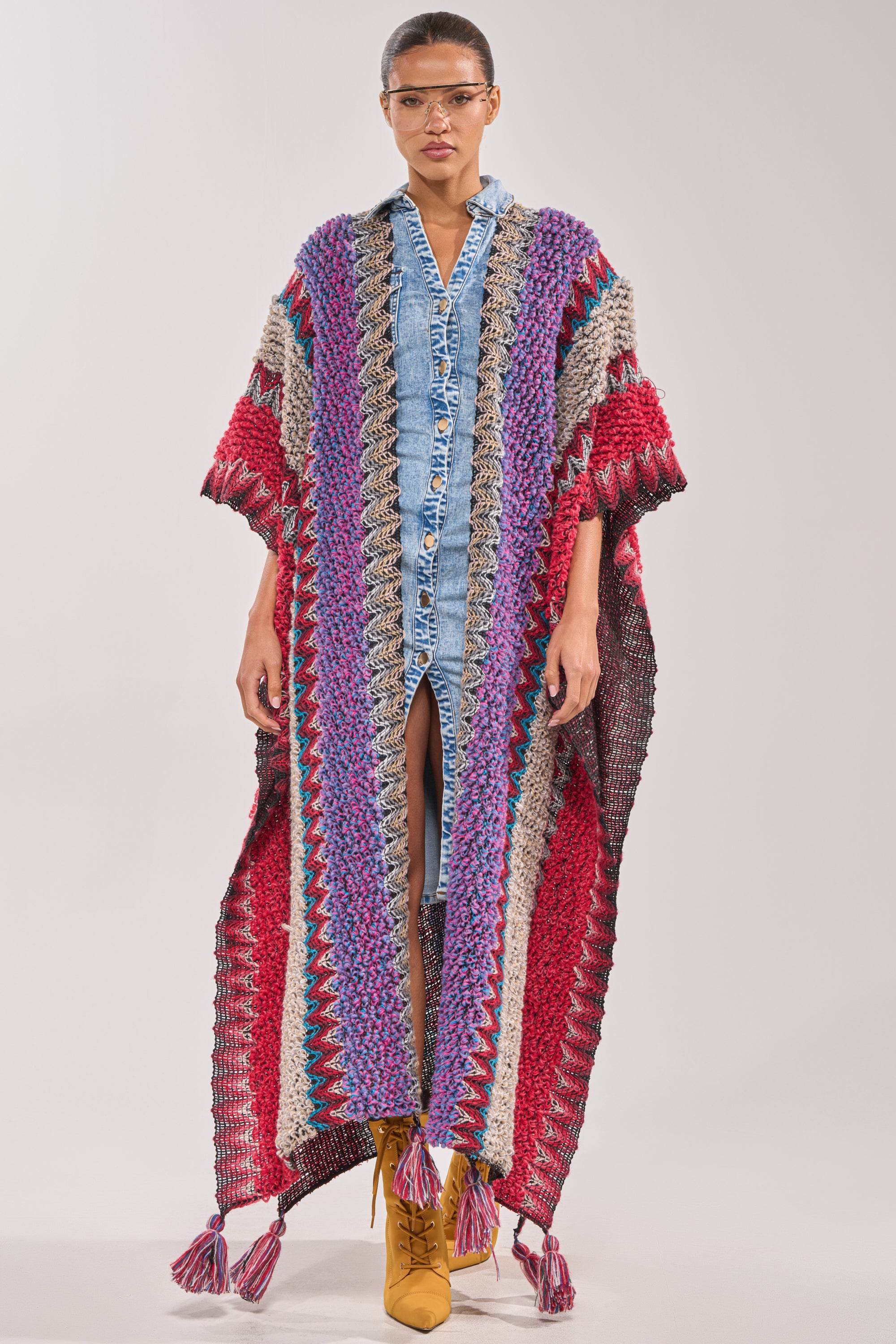 A person stands against a plain light background wearing the JUST WHAT I AM KIMONO—a colorful, striped boho kimono with tassels at the hem—layered over a blue denim button-down dress, paired with yellow boots and clear eyeglasses. 0