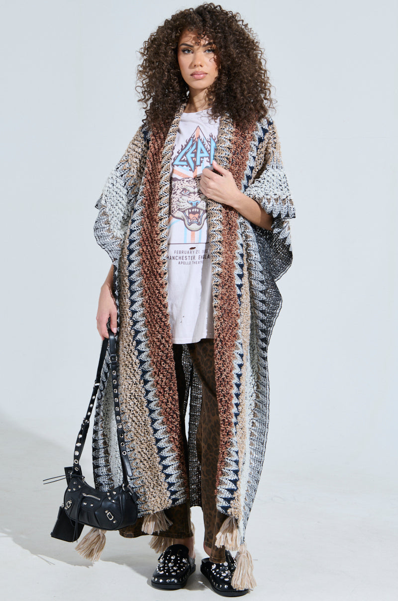 Wearing their curly hair with a hippie-chic flair, they sport the CRAZY FOR YOU CARDIGAN, a graphic T-shirt, and striped pants. Black studded shoes add edge as they confidently carry a black shoulder bag against a plain backdrop.