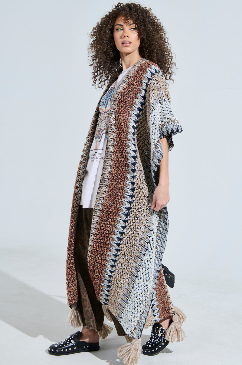 A person with curly hair models the CRAZY FOR YOU CARDIGAN, a colorful striped knit with fringe. They pair it with a white graphic tee, dark pants, and black loafers featuring silver accents while standing in a well-lit space.