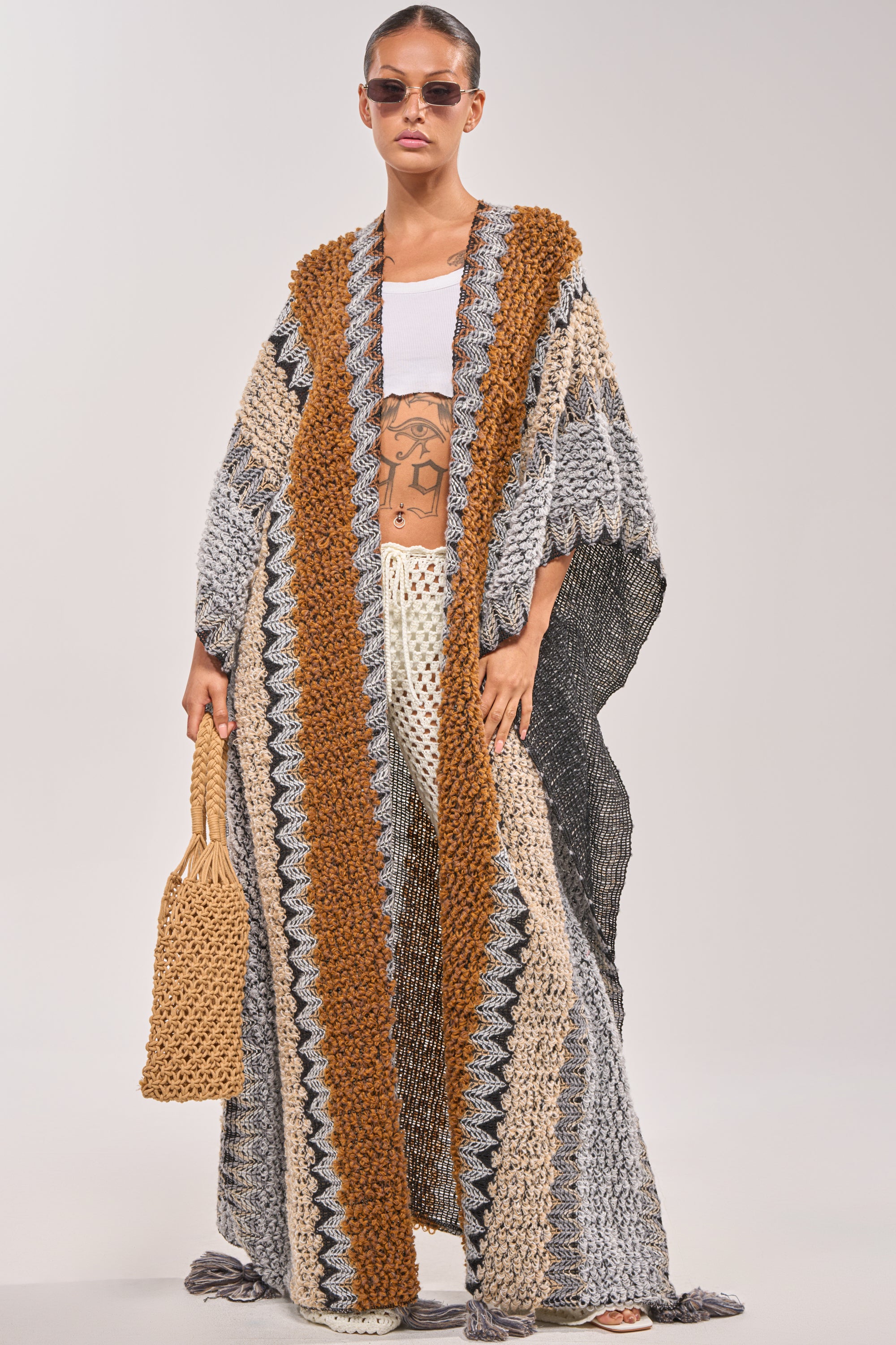 A person wearing sunglasses and the CRAZY FOR YOU CARDIGAN—a long, multicolored knit with gray, beige, and brown zigzag patterns—over a white top and crocheted pants, holding a woven bag. 1